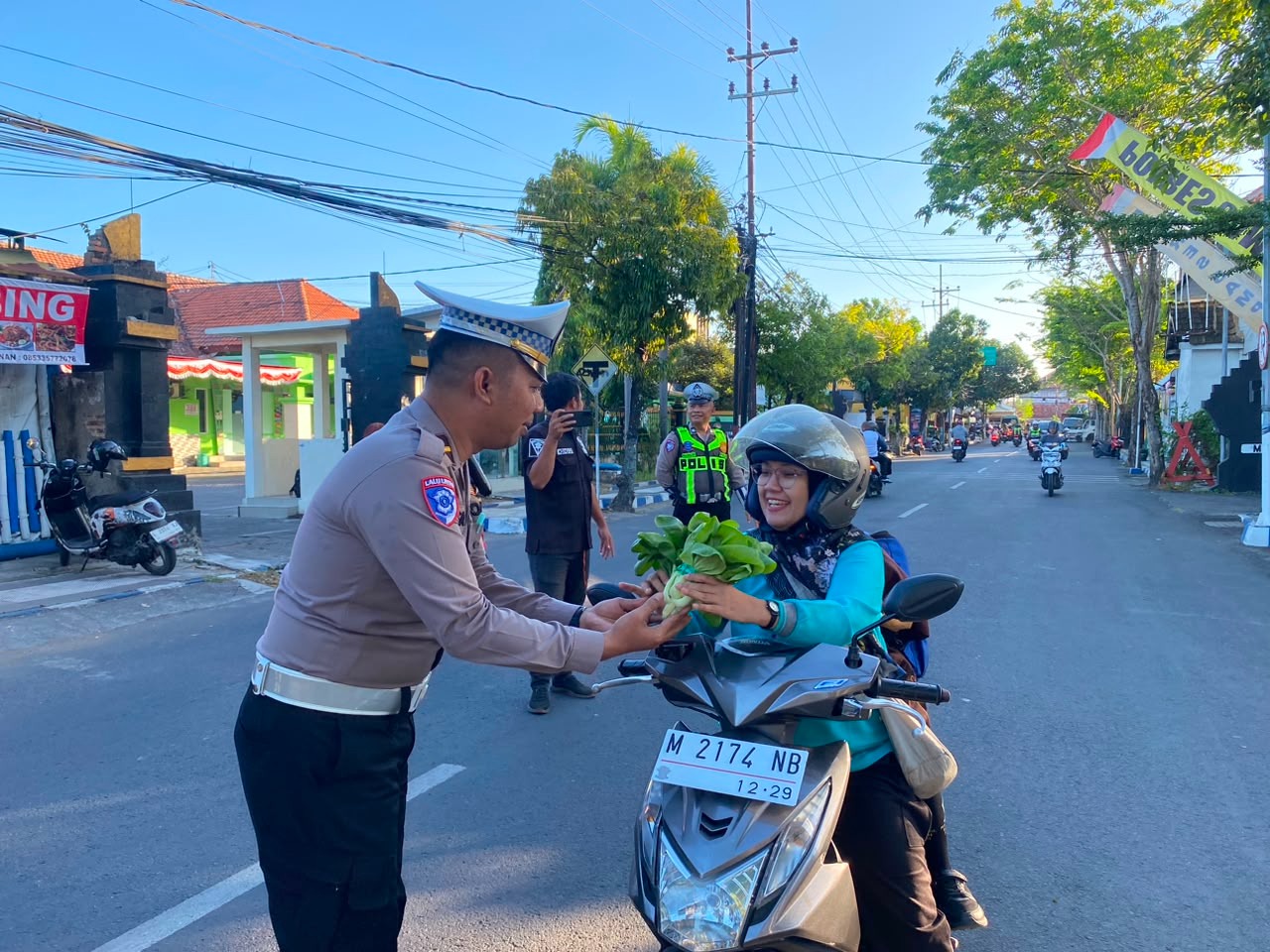 Caption Foto: Personel Satlantas Polres Sampang membagikan sayuran segar hasil Program Pekarangan Pangan Lestari (P2L) kepada pengguna jalan di sela pengaturan arus lalu lintas pada Jumat (24/7/2026) sebagai wujud pelayanan humanis sekaligus edukasi keselamatan berkendara.